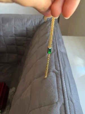 Guldhalsband med grön sten - Snyggt halsband i guldfärgad metall med en rektangulär grön sten som hänge. Kedjan är tunn och elegant, perfekt för att ge en färgklick till din outfit. Stenen har en djup smaragdgrön ton och infattningen är stilren och modern.