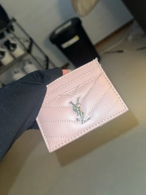 Rosa YSL kortfodral i läder - Säljer ett snyggt rosa kortfodral från YSL i äkta läder. Kortfodralet har silverfärgad YSL-logga framtill och dekorativa sömmar i chevronmönster. Perfekt för att hålla koll på dina kort med stil.