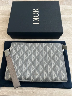 DlOR DIMOND MESSENGER POUCH - Säljer en fantastiskt fin Pouch i den ikoniska grå färgen med CD Diamond-mönstret. Väskan är en  klassiker och kombinerar lyx med praktisk funktion. Detaljer: Modell: CD Diamond Canvas & Smooth Calfskin. Färg: Grey (klassisk ljusgrå). Material: Slitstark belagd canvas med detaljer i mjukt kalvskinn Tillbehör: Kommer med originalbox, dustbag och den avtagbara axelremmen. Skickas spårbart och försäkrat.Tveka inte att höra av dig vid frågor eller om du vill ha fler bilder!