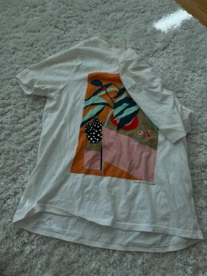 Vit t-shirt med färgglatt tryck - Säljer en vit t-shirt med ett stort färgglatt grafiskt tryck på ryggen. Trycket har inslag av orange, rosa, grön och svart med motiv av växter och en prickig vas. T-shirten har korta ärmar och avslappnad passform. Perfekt för dig som gillar unika prints. Storlek Xs men är en mer en S