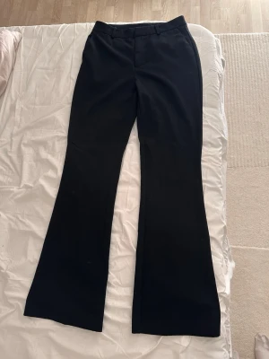 Svarta utsvängda kostymbyxor - Säljer ett par svarta kostymbyxor med utsvängda ben och hög midja. Byxorna har hällor för skärp och klassisk dragkedja framtill. Perfekta för en stilren och trendig look. Materialet känns mjukt och följsamt, troligen polyesterblandning.