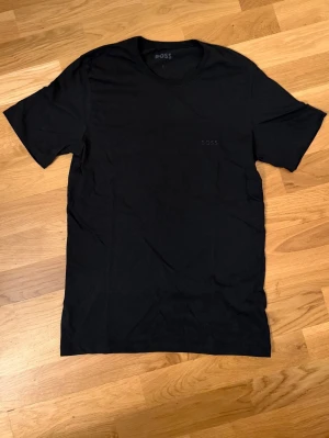 Svart t-shirt från BOSS - Snygg svart t-shirt från BOSS med diskret logga på bröstet. Klassisk rund halsringning och korta ärmar. Tillverkad i mjukt material som känns skönt mot huden. Perfekt basplagg för en clean och stilren look. T-shirten är slim fit. Pris kan diskuteras 