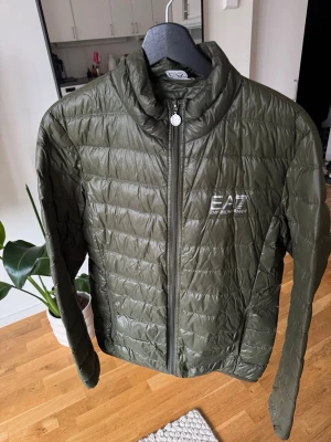 Olivgrön dunjacka från Emporio Armani - Snygg olivgrön dunjacka från Emporio Armani EA7 med quiltad design och hög krage. Jackan har dragkedja framtill och logga på bröstet. Perfekt för kalla vinterdagar och ger en stilren look.