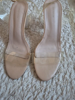 Beige transparenta pumps från Shein - Säljer ett par snygga pumps från Shein i beige med transparent rem över tårna. Skorna har en öppen tå och klack, samt en stilren design med plastdetaljer som ger en modern look.
