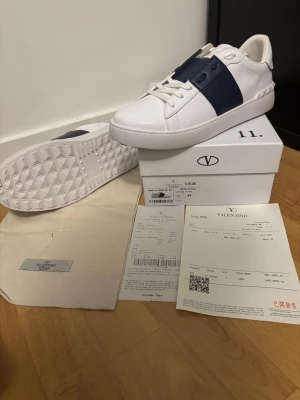 Valentino vita sneakers med blå detalj - Snygga vita sneakers från Valentino med bred mörkblå rem över mitten och vita skosnören. Skorna har en klassisk låg profil och är tillverkade i slätt läder med gummisula. Perfekta för dig som vill ha en stilren och exklusiv look.