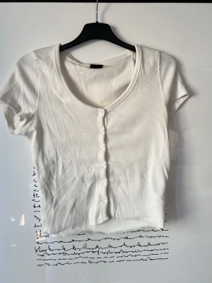 Vit ribbad topp med knappar - Kortärmad, croppad topp i vit ribbad bomull med rund halsringning och dekorativa knappar framtill. Perfekt till jeans eller kjol för en enkel och clean look. Pris kan diskuteras.