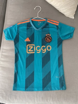 Ajax bortatröja Adidas turkos - Snygg turkos Ajax bortatröja från Adidas med orange detaljer och Ziggo-logga på bröstet. Tröjan har svarta diagonala ränder, korta ärmar och klassiska Adidas-ränder på axlarna. Tillverkad i lätt och ventilerande material, perfekt för träning eller match.