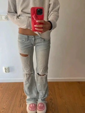 Ljusblå slitna jeans med hål - Säljer ett par ljusblå jeans med raka/bootcut ben och slitna detaljer, inklusive hål på båda knäna. Jeansen har låg midja. Perfekta för en avslappnad och trendig look.