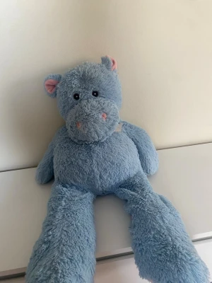 Blått gosedjur flodhäst med rosett - Mysig blå flodhäst som gosedjur med långa ben och små rosa detaljer på nos och öron. Den har en söt ljusrosa rosett runt halsen och är tillverkad i mjuk plysch. Perfekt att krama eller ha som dekoration i rummet.