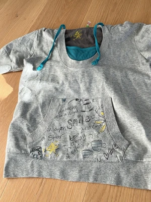 Grå hoodie med doodle-tryck - Grå hoodie med turkosa dragsnören och stor magficka. Fickan har handritade doodles och texter i svart, blått och gult, bland annat tekoppar, stjärnor och ord som 'smile'. Hoodien har huva och är gjord i mjukt sweatshirtmaterial. Hoodien är kortärmad. På lappen står det strl 38/40 men är som en 34/xs. På en bild ser ni lappen i tröjan men jag fattar inte vilket märke det är.