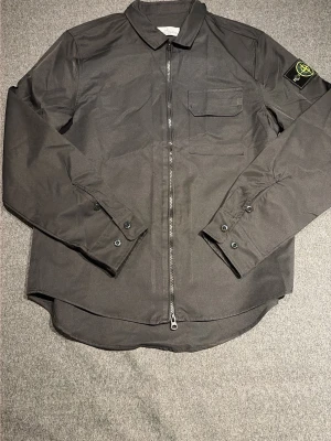 Svart overshirt från Stone Island - Snygg svart overshirt från Stone Island med dragkedja framtill, klassisk krage och bröstficka. Ikonisk patch med logga på vänster ärm. Tillverkad i ett slitstarkt material som ger en clean och modern look. Priset är inte hugget i sten