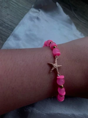 Rosa pärlarmband med sjöstjärna - Snyggt armband med starkt rosa pärlor och små guldfärgade detaljer. Armbandet har en guldfärgad sjöstjärneberlock som ger en somrig och lekfull vibe. Perfekt accessoar för att liva upp din outfit.