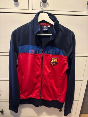 Vintage FC Barcelona träningsjacka blå/röd - Snygg träningsjacka från FC Barcelona i blått och rött med klubbens logga på bröstet. Jackan har hel dragkedja, hög krage och två fickor med dragkedja. Perfekt för dig som älskar fotboll och vill representera ditt favoritlag.
