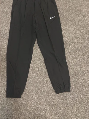 Svarta träningsbyxor från Nike - Säljer ett par svarta träningsbyxor från Nike med resår i midjan och vid bensluten. Byxorna har en enkel design med en vit Nike-logga på vänster lår. Perfekta för gymmet eller löprundan.