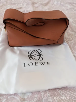 Brun Puzzle hobo bag från Loewe - Säljer en brun Puzzle Bag från Loewe i slätt läder. Väskan har en unik geometrisk form, bred axelrem och guldfärgad dragkedja. Loewe-loggan är präglad på framsidan och dustbag medföljer. Perfekt för dig som gillar stilrena och exklusiva accessoarer.