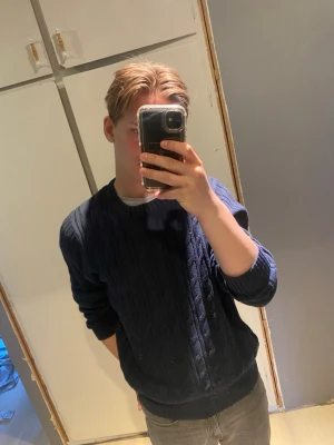 Race marine tröja - Säljer en mörkblå kabelstickad tröja med rund hals och ribbade muddar. Tröjan har ett diskret märke nertill och är perfekt för dig som gillar klassisk stil. Materialet känns mjukt och bekvämt, och modellen är normal i passformen.