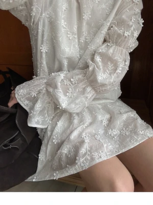 Vit blommig kortklänning med öppen rygg - Supersöt vit kortklänning med broderade blommor, volangkant och långa puffärmar. Klänningen har öppen rygg med knytband upptill och är gjord i ett tunt, lätt material som känns luftigt. Perfekt för sommarens alla tillfällen! Använd 1 gång. 