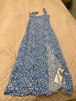 Blåvit blommig långklänning - Söt långklänning med blått och vitt blommönster, smala axelband och knyt fram. Klänningen har en slits på sidan och är gjord i ett lätt och luftigt material, perfekt för varma dagar.