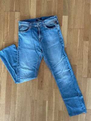 Slim Replay tchad jeans - Säljer ett par klassiska blå jeans från Wrangler med raka ben och fem fickor. Jeansen har snygga slitningar och ljus tvätt, samt detaljerade sömmar på bakfickorna. Nypris: 900kr
