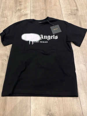 Svart Palm Angels Las Vegas t-shirt - Svart t-shirt från Palm Angels med tryck i vitt framtill där det står 'Angels Las Vegas' och en vit spray-effekt över loggan. Klassisk rund hals och korta ärmar. Perfekt för dig som gillar streetwear och vill sticka ut.
