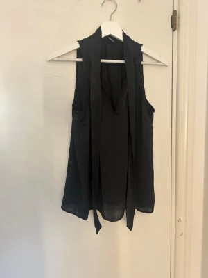 Svart ärmlös blus med knytband - Snygg svart ärmlös blus med långa knytband i halsen som kan knytas till en rosett bak. Blusen har en lätt v-ringning framtill och är gjord i ett tunt, flygigt material som ger ett elegant intryck.