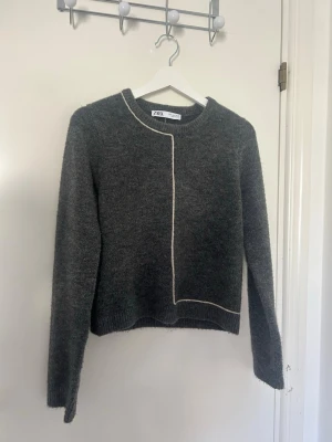 Mörkgrå stickad tröja från Zara - Mysig mörkgrå stickad tröja från Zara med lång ärm och rund hals. Tröjan har en unik vit kontrastsöm som går längs framsidan och nederkanten, vilket ger en cool och modern look. Perfekt till höst och vinter för en stilren outfit.