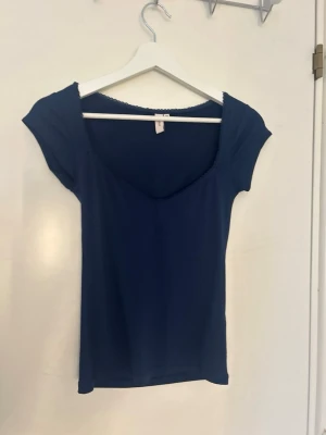 Mörkblå topp från Nelly med spetskant - Mörkblå topp från Nelly med söt spetskant runt den djupa v-ringningen. Kortärmad och figurnära modell i mjukt material, perfekt för att styla med jeans eller kjol. Enkel men ändå feminin design.