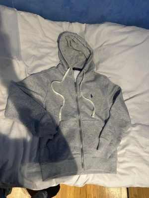 Grå  hoodie från Polo Ralph Lauren - Snygg grå zip hoodie från Polo Ralph Lauren med vit dragsko i huvan och klassisk blå logga på bröstet. Tillverkad i mjukt sweatshirtmaterial, har fickor framtill och ribbade muddar vid ärmar och nederkant. Perfekt för en chill och stilren look.