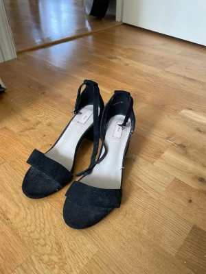 Svarta klackskor  - Stilrena svarta klackskor med öppen tå. Skorna har ett skysterbart ankelband. Materialet är mockaliknande och de är bara använda en gång.