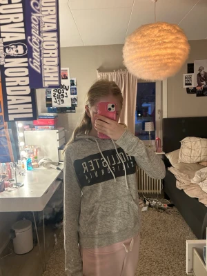 Grå Hollister hoodie med tryck - Säljer en grå hoodie från Hollister med stort vitt tryck på bröstet där det står 'Hollister California'. Hoodien har huva med vita snören och är tillverkad i mjukt sweatshirtmaterial. Perfekt för en avslappnad och trendig look.