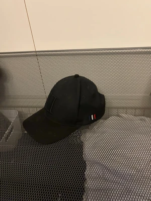 Svart keps från Les Deux - Snygg svart keps från Tommy Hilfiger med böjd skärm och diskret logga framtill. På sidan finns de klassiska Hilfiger-färgerna i rött, vitt och blått. Kepsen är i bomull och har justerbar rem bak för perfekt passform.