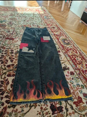 Svarta jeans med flames och stjärnor - Unika svarta jeans med bred passform, dekorerade med handmålade flames i rött, orange och gult längst ner på benen. Framtill finns färgglada tygpatches och en svart stjärna, baktill två stora vita stjärnor. Perfekta för dig som vill sticka ut med din stil.