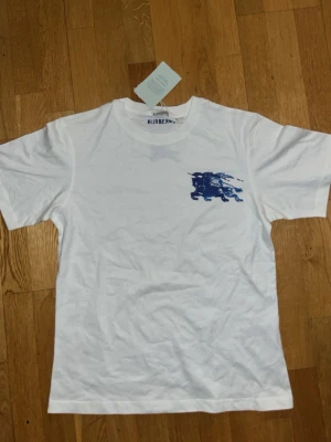 Vit Burberry t-shirt med blått tryck - Säljer en vit t-shirt från Burberry med ett blått tryck av en riddare på bröstet. Klassisk rund hals och korta ärmar. Tillverkad i mjuk bomull för en skön känsla och stilren look. Perfekt för dig som gillar exklusiva märken och enkel design.