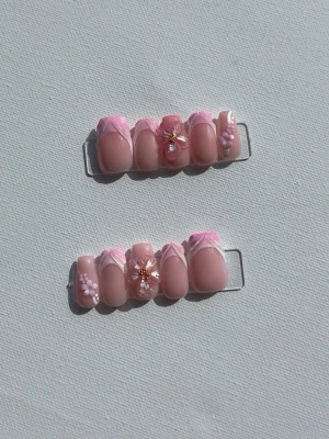 Press-on naglar rosa 3D-blommor - Säljer ett set press-on naglar i ljusrosa med vita och rosa franska tippar. Naglarna har 3D-blommor, pärldekor och guldglitterdetaljer. Formen är oval och materialet är gel. Perfekt för dig som vill ha en söt och unik nageldesign.
