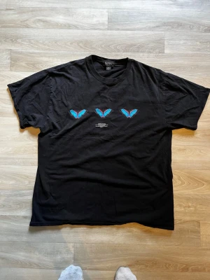 Svart t-shirt med fjärilar från MAN - Svart t-shirt från MAN med tre blå fjärilar tryckta på bröstet och stora fjärilar samt texten 'official man' på ryggen. T-shirten har rund hals, korta ärmar och en relaxed fit. Perfekt för dig som gillar streetwear och coola prints.