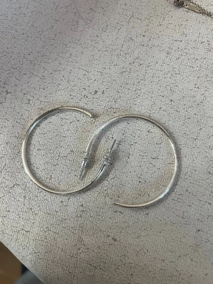Stora silvriga hoops örhängen - Säljer ett par stora, runda hoops i silverfärgad metall. Örhängena har en enkel och stilren design utan dekorationer, perfekta för dig som gillar minimalistiska accessoarer. Passar till många olika stilar och outfits.