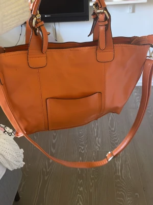 DavidJones väska  - Snygg orange-brun väska i slätt skinn med både handtag och justerbar axelrem. Väskan har en enkel, stilren design med en ficka på framsidan och guldfärgade metalldetaljer. Perfekt för dig som vill ha en färgklick till din outfit.