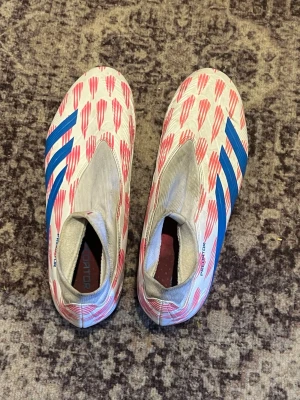 Adidas Predator fotbollsskor - Säljer ett par Adidas Predator fotbollsskor i vitt med röda detaljer och blåa ränder. Skorna har en strömlinjeformad design utan snörning och är tillverkade i syntetmaterial för bästa bollkontroll. Perfekta för dig som vill sticka ut på planen.