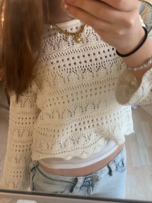 Vit virkad stickad tröja - Säljer en vit virkad stickad tröja med lång ärm och vågig kant nertill. Tröjan har ett detaljerat hålmönster och är lätt genomskinlig, perfekt att styla med ett linne under. Passar till jeans för en avslappnad men trendig look.