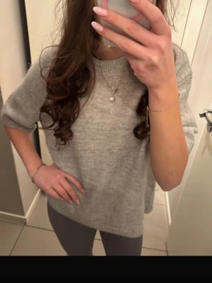 Grå stickad tröja med kort ärm - Mysig grå stickad tröja med korta ärmar och rund hals. Tröjan har en avslappnad passform och ribbstickade kanter. Perfekt att matcha med jeans eller kjol för en enkel och stilren look. Passar både vid kyligare temperaturer och när solen är framme ☀️