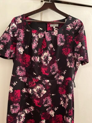 Blommig klänning från Debenhams - Snygg blommig klänning från Debenhams med korta ärmar och rundad halsringning. Klänningen har ett mönster i rosa, rött och vitt på svart botten och är figurnära med veckad detalj vid halsen. Perfekt för dig som vill sticka ut med färg och mönster.