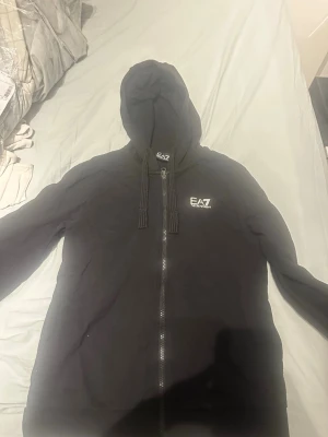 Svart EA7 zip hoodie - Säljer en svart zip hoodie från EA7 Emporio Armani. Hoodien har dragkedja framtill, justerbar huva med snören och en liten EA7-logga på bröstet. Perfekt för en chill och stilren look. Materialet känns mjukt och bekvämt.
