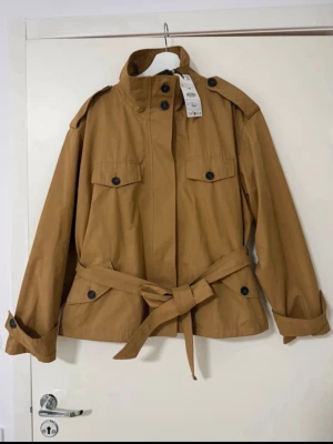 Kamelbrun trenchcoat med bälte - Stilren kamelbrun trenchcoat med knytskärp i midjan, axelklaffar och svarta knappar. Två bröstfickor med lock och knapp samt dekorativa detaljer vid ärmslut. Klassisk krage och ventilerad rygg. Perfekt för en trendig och avslappnad look.