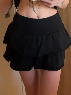 Svart volangkjol med bred midja - Supersöt svart kortkjol med dubbla volanger och bred midjeresår. Kjolen har en flowy look och passar perfekt till crop tops eller stickade tröjor. Tillverkad i mjukt tyg som ger en skön känsla och snygg siluett.