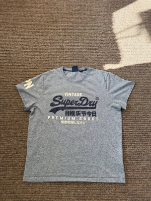 Grå Superdry t-shirt  - Passar XL. Snygg grå t-shirt från Superdry med stort vintage-tryck i svart och vitt på bröstet samt textdetalj på ärmen. Klassisk rund halsringning och normal passform. Perfekt för en avslappnad och trendig stil.