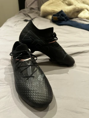 Svarta Puma Future fotbollsskor - Säljer ett par svarta Puma Future fotbollsskor med låg profil och stickad strumpliknande krage. Skorna har snörning, diskret mönster på ovandelen och Puma-logga i rosa på sidan. Tillverkade i syntetmaterial för bra grepp och kontroll på planen. Nypris 1200kr endast testade :) 