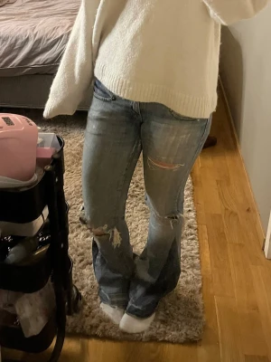 Blå slitna bootcut jeans - Säljer ett par blå bootcut jeans med slitningar och hål på knäna. Jeansen har en klassisk femficksmodell och är tillverkade i denim med en tvättad look. Perfekta för en avslappnad och trendig stil.