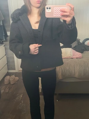 Svart ullkappa med fuskpäls - Kort svart ullkappa med bred krage och fuskpälsdetalj. Kappan har stora knappar vid ärmsluten och en rak, boxig passform. Perfekt för dig som vill ha en stilren och trendig look till hösten eller vintern.