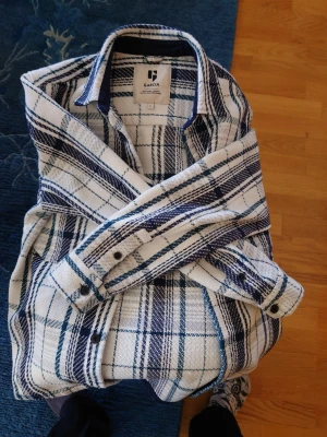 Vit och blå rutig overshirt Garcia - Overshirt från Garcia i vit och blå rutig design. Skjortjackan har klassisk krage, knäppning framtill och på ärmsluten samt två bröstfickor. Materialet är grovt och ger en avslappnad streetkänsla. Perfekt lager-på-lager-plagg för kyligare dagar.