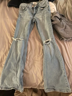 Ljusblå utsvängda jeans med hål - Säljer ett par ljusblå jeans från 157 med utsvängda ben och slitna hål vid knäna. Jeansen har klassisk femficksdesign och hög midja. Perfekta för en avslappnad och trendig look. Storlek S short